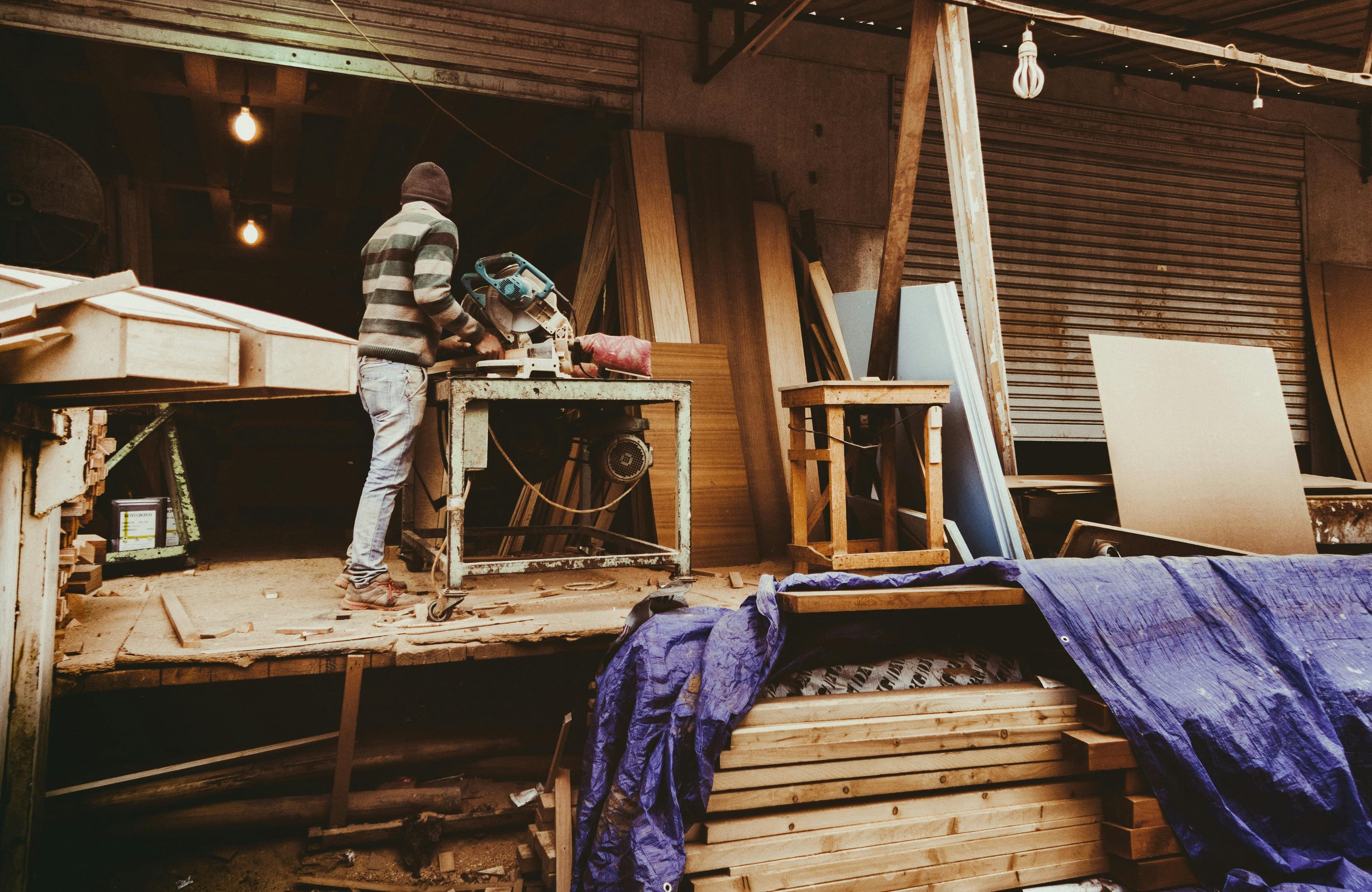 Carpenter in striped sweater cutting wood with a miter saw in a busy, rustic workshop.